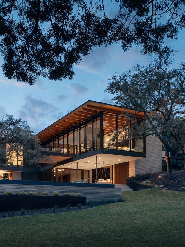 A contemporary two-storey residence viewed from the landscaped front garden at dusk. The home has a dramatic pitched roof with deep exposed timber eaves, extensive floor-to-ceiling glazing, dark slender framing, warm timber wall cladding, and a projecting upper-level volume. Steps, terraced retaining walls, lawn, and mature trees surround the building, while warm interior lighting creates an inviting glow through the glass.