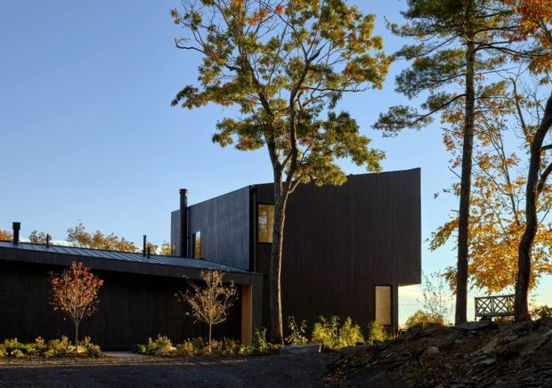 Exterior view of a dark, minimalist modern residence set among mature deciduous trees on a gently sloped, landscaped site. The building has a strong rectilinear form, low-pitched rooflines, dark vertical cladding, tall narrow windows, and a small deck or railing visible at the right.