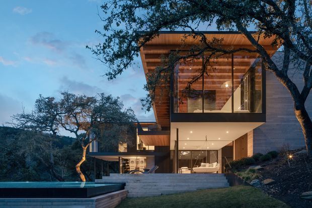 Exterior view of a contemporary multi-level residence set into a wooded, sloping site at dusk. The house features extensive floor-to-ceiling glazing, projecting upper volumes, warm timber soffits and exposed timber structure, dark horizontal cladding, broad concrete steps, and a long reflecting or swimming pool in the foreground. Interior living spaces are visible through the glass.