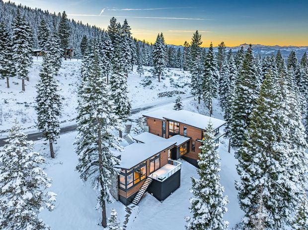 A modern multi-level timber-clad house with expansive glazing sits among snow-covered evergreen trees on a mountainous, wooded site. Flat or very low-pitched roof planes, dark window framing, and warm interior lighting contrast with the white snow, while a winding road passes behind the property and distant mountain ridges are visible at sunset.