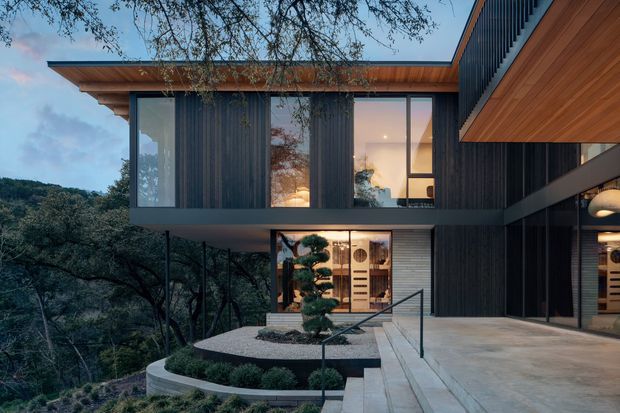 Contemporary multi-level residential house set into a wooded, sloping landscape. The facade combines extensive floor-to-ceiling glazing, dark vertical cladding, warm timber soffits and wall lining, and crisp cantilevered horizontal forms. A landscaped entry terrace with a sculpted tree, gravel garden and streamlined stair railing leads toward the glazed interior.