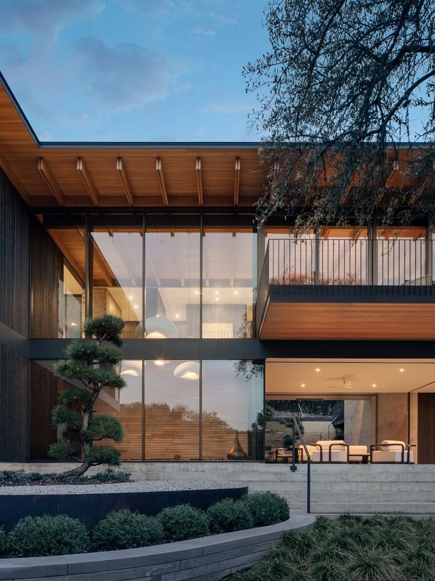 Exterior architectural view of a contemporary multi-level residence with extensive floor-to-ceiling glazing, dark vertical cladding, warm timber soffits and balcony cladding, exposed roof overhangs, and an open ground-level living area. Concrete steps, landscaped garden beds, and sculptural planting lead toward the house. The interior is visible through the glazing, including pendant lighting and lounge furnishings.