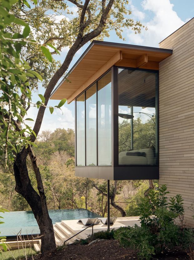 Exterior architectural view of a contemporary home set among mature trees, featuring a projecting glazed room with dark window frames, a warm timber-lined soffit, and light brick wall cladding. A swimming pool and terrace are visible below, with a natural wooded landscape beyond.