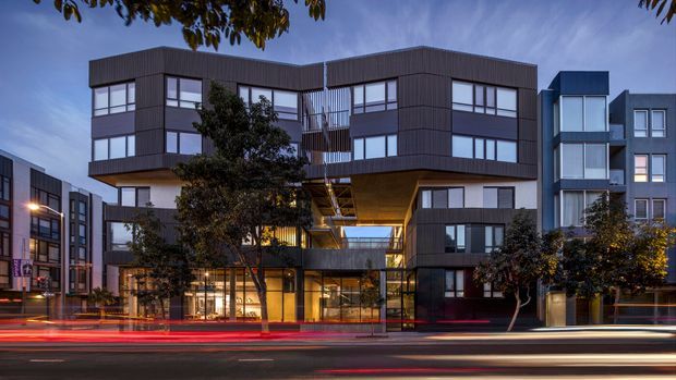 Twilight exterior view of a contemporary multi-storey urban apartment or mixed-use building. The facade combines dark vertical cladding, extensive rectangular glazing, projecting box-like volumes, recessed balconies, and a dramatic illuminated cantilevered central section. Adjacent modern buildings and light trails from passing traffic reinforce the dense urban setting.