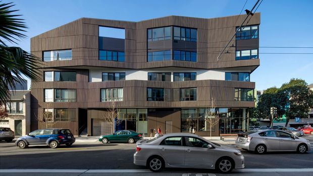 Street-level exterior view of a contemporary multi-storey urban building with projecting and recessed volumes, extensive dark-framed glazing, and a vertical timber-look cladding facade. Commercial or mixed-use activity is visible at ground level, with apartments or offices above. The building fronts a busy city street with parked and passing cars, overhead utility wires, and limited surrounding landscaping.