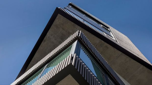 Low-angle exterior view looking up at a tall contemporary building. The facade features projecting concrete slabs, dark structural bands, extensive reflective glazing, and dense vertical ribbed fins wrapping the corners and upper levels against a clear blue sky.
