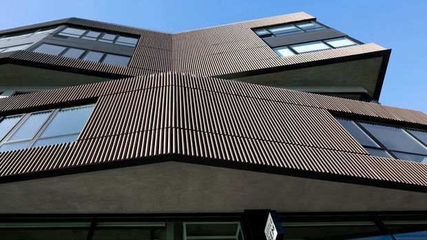 Upward-looking view of a multi-storey contemporary building facade with projecting angular volumes, extensive dark-framed glazing, and a distinctive vertical ribbed exterior screen or cladding. The facade is predominantly dark grey and brown under a clear blue sky, with strong geometric lines and deep overhangs.