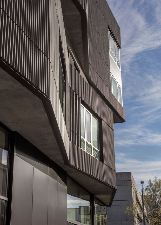 Vertical exterior view of a contemporary multi-storey building with dark ribbed facade cladding, projecting concrete slabs and balconies, tall glazed windows, and a ground-level storefront entrance. The image emphasizes the building facade and strong geometric composition against a blue sky and urban streetscape.