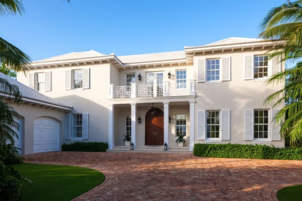 A large, symmetrical two-storey pale stucco residence with a low-pitched light roof, white shutters, classical columns, decorative balcony railings, and a prominent timber entry door. The home is approached by a curved brick-paved driveway and framed by manicured hedges, lawn, and tropical palm trees.