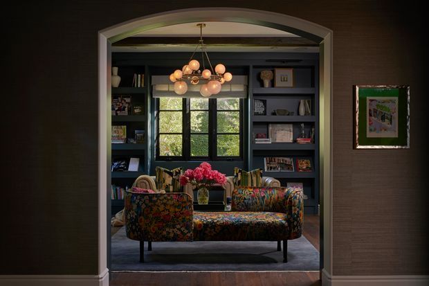 A dark, richly styled sitting room or library viewed through a wide arched opening. Built-in charcoal shelving flanks a black-framed window, while a colourful patterned sofa and matching armchairs sit beneath a sculptural globe pendant light. The room has dark painted walls, timber flooring, a large rug, layered decorative objects, and a framed artwork on the adjacent wall.