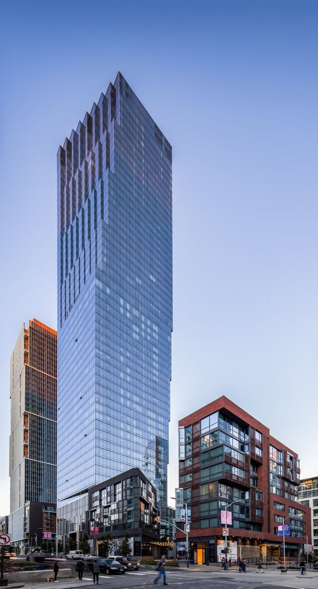 Urban streetscape photograph featuring a tall, sharply angular glass office tower as the dominant subject, with adjacent mid-rise brick-and-glass apartment or commercial buildings, street-level retail, roads, and pedestrians. The architecture has a contemporary high-rise character with extensive blue-tinted curtain-wall glazing and prominent vertical geometric fins.