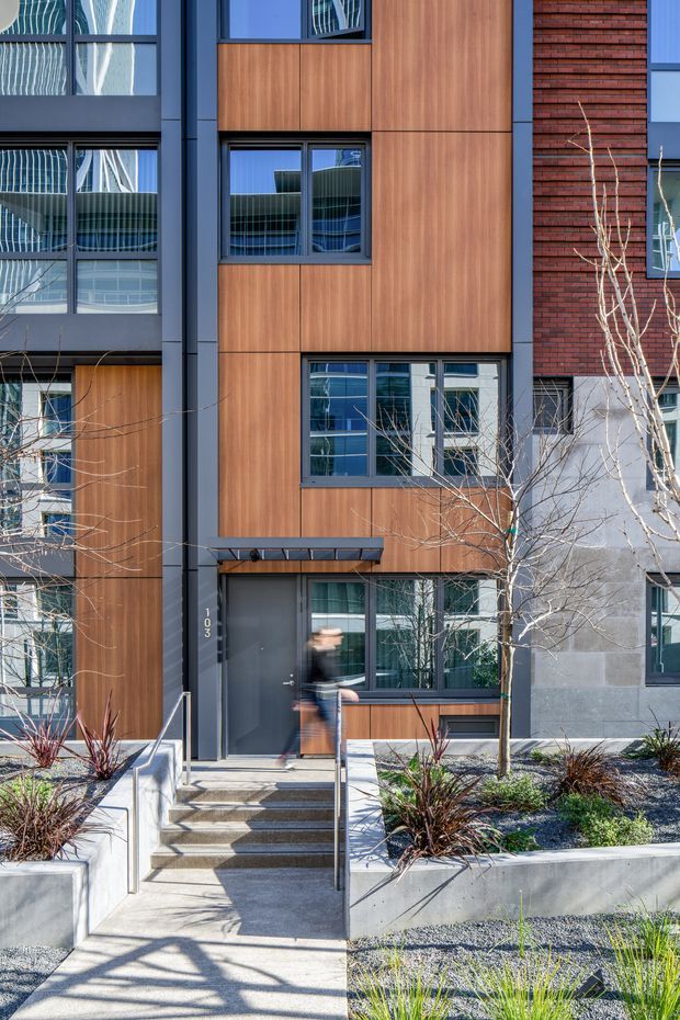 Street-level view of a contemporary multi-storey residential building facade, featuring warm wood-look cladding, dark metal window and door frames, red brickwork, extensive glazing, and a landscaped concrete entry with stairs and a narrow canopy. The facade is shown in bright daylight with reflections of neighboring modern buildings in the windows.