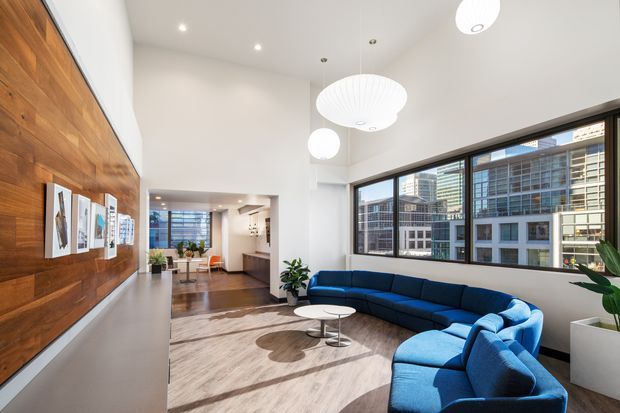 Bright contemporary living room with a large blue sectional sofa, round coffee tables, pale area rug, white walls, and sculptural pendant lights. A full-height warm timber feature wall with framed artwork lines the left side, while expansive black-framed windows overlook dense urban buildings. The open-plan space extends toward a dining area and kitchen beyond.