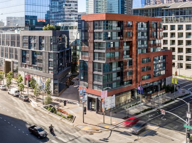 Elevated urban view of a dense city intersection surrounded by mid- and high-rise commercial and residential buildings. The main foreground building has a contemporary red-brick facade with extensive floor-to-ceiling glazing, dark metal framing, projecting volumes, and horizontal louvred screens. Adjacent buildings use glass curtain walls, dark cladding, and light masonry. Streets, sidewalks, traffic signals, and landscaped street trees are visible below.