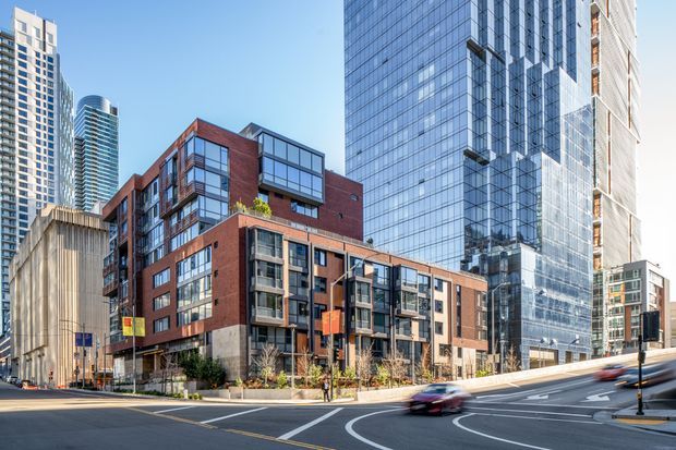 Street-level urban architectural view showing a contemporary mid-rise residential or mixed-use brick-and-glass building in the foreground, surrounded by taller glass and concrete high-rise towers. The scene includes landscaped sidewalk planting, multilane roads, traffic markings, and dense downtown development under clear daylight.