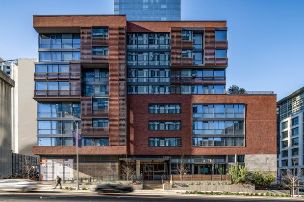 Exterior street-level photograph of a multi-storey urban building with a contemporary composition of red-brown brick, horizontal timber-toned screening, extensive blue-grey glazing, projecting balconies, and a recessed central entrance. The building sits within a dense city streetscape with neighbouring commercial or residential buildings visible around it.