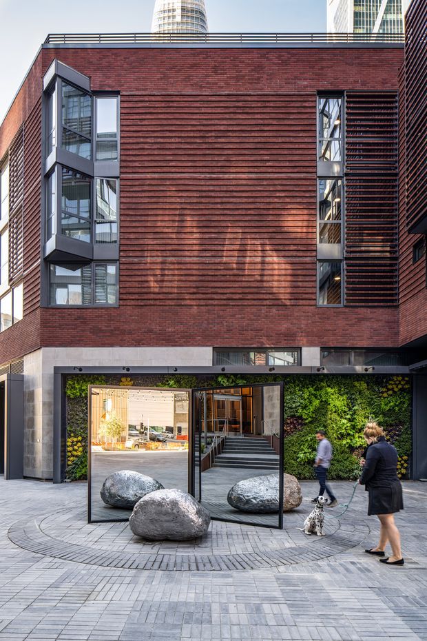 Urban exterior view of a contemporary multi-storey building with a dark red brick facade, horizontal brick detailing, projecting glazed bay windows, narrow vertical windows with external louvres, and a rooftop railing. The ground-level entrance is framed by grey stone or concrete and a lush planted wall, with large freestanding boulders and a paved public forecourt. High-rise buildings are visible in the background.