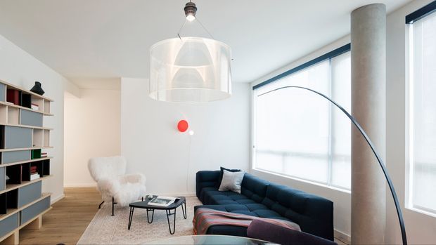 Bright contemporary living room with large corner windows, a dark navy sectional sofa, sculptural arched floor lamp, white pendant light, round concrete column, light timber flooring, pale area rug, and built-in open shelving along the left wall. A white furry accent chair and small black coffee table complete the seating area.