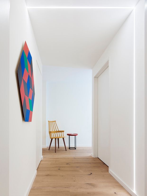 Bright, minimalist hallway with white walls and ceiling, a warm light-oak timber floor, and concealed linear lighting along the right wall and ceiling edge. A colourful geometric artwork hangs on the left wall, while the corridor opens to a small seating area with a timber spindle-back chair and a compact red-top stool. White flush doors and crisp architectural lines create a clean, contemporary feel.
