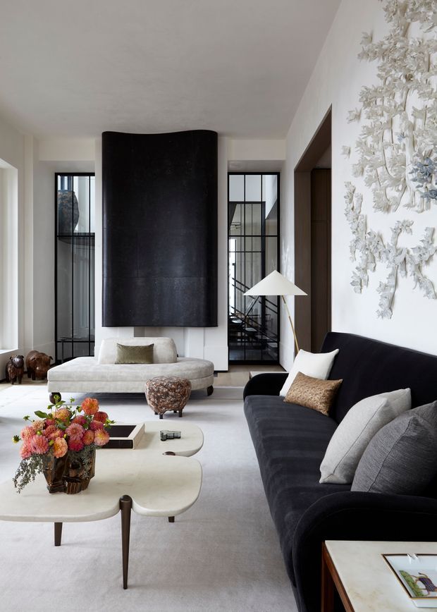 Refined contemporary living room with a pale neutral palette, large black curved feature fireplace or chimney breast, black-framed internal glazing, a dark velvet sofa with light cushions, sculptural cream coffee tables, and a prominent white textured floral wall artwork. A staircase or circulation space is visible through the glazed partitions.