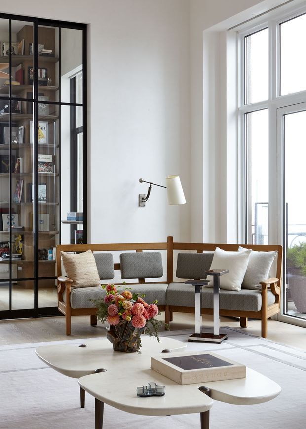 Bright, refined living room with a mid-century-inspired timber sofa, upholstered grey cushions, sculptural white coffee tables, a slim wall-mounted reading lamp, and a large floral arrangement. A black-framed glazed built-in shelving unit displays books and objects along the left wall, while tall white-framed windows provide abundant natural light and views outside. The room has a calm, curated contemporary character with warm timber accents.