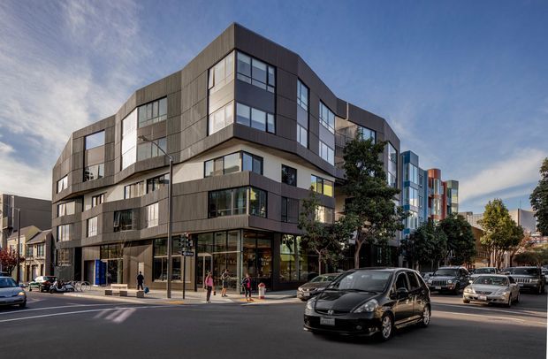 Street-level view of a contemporary multi-storey urban apartment or mixed-use building on a city corner. The building features stepped and curved massing, dark vertical metal cladding, extensive glazing, projecting balconies, and ground-floor commercial-style glazing. Adjacent modern buildings, street trees, pedestrians, and traffic reinforce the dense urban setting.