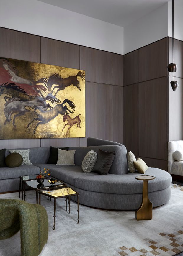 A refined contemporary living room with a large curved grey sectional sofa, dark wood wall panelling and integrated cabinetry, a dramatic equestrian artwork, sculptural pendant lights, glass-and-metal coffee tables, and a neutral textured rug. The room has a sophisticated, moody palette with warm metallic accents.