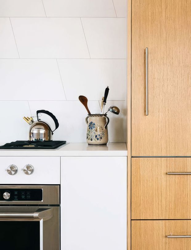 Close-up view of a contemporary kitchen with white slab cabinetry and large white tiled splashback, a black cooktop and stainless-steel oven, and warm light-oak pantry cabinetry with long brushed metal handles. A kettle and decorative utensil crock sit on the benchtop, adding a subtle lived-in detail.