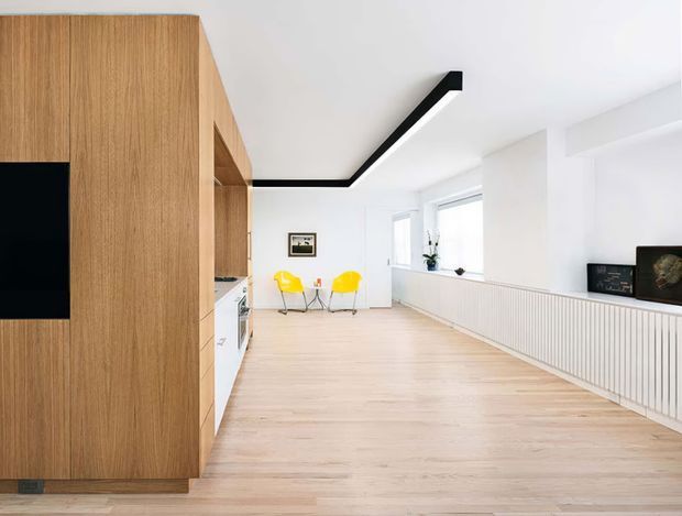 Bright, minimalist open-plan interior with pale timber flooring and a full-height timber joinery wall incorporating a black television panel and a partially visible kitchen. White walls and cabinetry, a long ribbed white built-in radiator or storage element beneath the windows, sculptural yellow chairs, and a bold linear black ceiling light create a clean contemporary composition.
