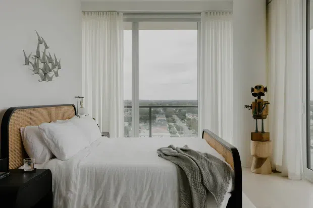 A bright, minimalist bedroom with a rattan-panel bed, white bedding, a soft grey throw, and a black bedside table. Full-height windows with sheer vertical blinds provide expansive city views, while sculptural wall art and a wooden decorative figure add understated character.