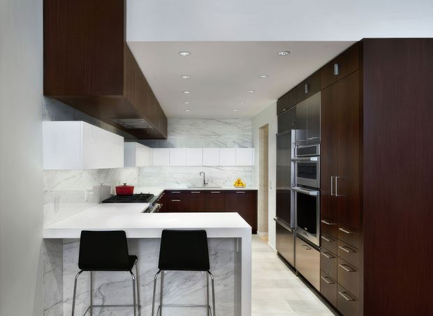 A sleek contemporary galley kitchen with a white waterfall-edge island and two black bar stools. Dark timber cabinetry runs along both sides, incorporating double ovens and integrated storage, while white marble-look wall surfaces and flooring create a bright, refined contrast. Recessed ceiling lights provide even illumination.