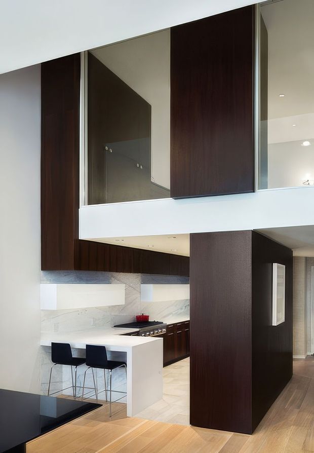 Contemporary open-plan kitchen and living area with a dramatic double-height volume. The space features dark timber wall panelling and cabinetry, white stone or painted surfaces, a compact white kitchen island with two stools, integrated stainless-steel appliances, and a mezzanine or upper-level walkway with clear glazing. Pale timber flooring and strong geometric architectural forms create a refined, high-contrast interior.