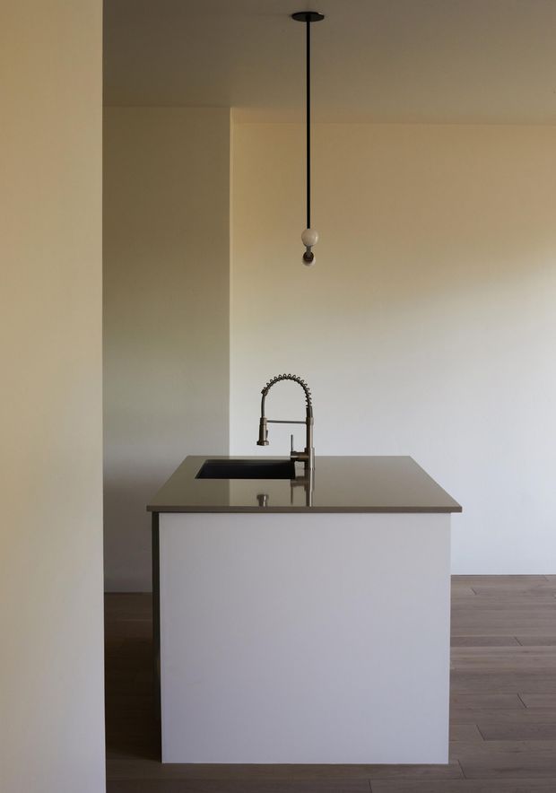 Minimalist kitchen interior viewed from an adjacent space, centered on a simple island with an integrated dark sink and tall gooseneck tap. The island has a plain white waterfall-style base and a dark, smooth benchtop. Warm timber flooring, off-white walls, and a single suspended pendant contribute to the sparse, quiet composition.