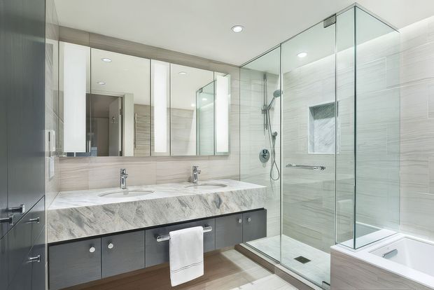 A bright, contemporary bathroom featuring a double vanity with dark built-in cabinetry and a pale stone benchtop, mirrored wall cabinets, a frameless glass walk-in shower, and a partially visible built-in bathtub. Light horizontally textured wall tiles and warm timber-look flooring create a clean, refined finish.