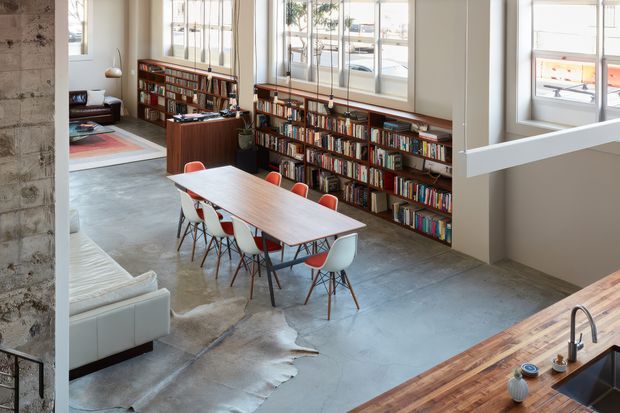 Open-plan industrial-style interior combining a dining area, living area, and kitchen edge. A long timber dining table with colourful molded chairs sits on a polished concrete floor, backed by extensive built-in bookshelves beneath tall industrial-style divided windows. A partial kitchen benchtop and sink appear in the foreground, while exposed concrete and timber elements reinforce the loft-like character.