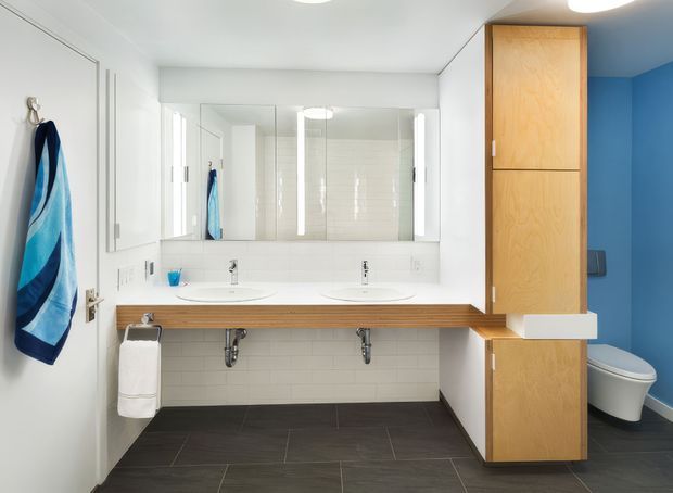 Bright contemporary bathroom with a wide double vanity, white countertop and integrated basins, large mirrored wall, white subway-tile wall finish, dark tiled floor, and warm timber cabinetry. A blue accent wall and partially screened toilet are visible at right. The room has clean-lined built-in joinery and modern ceiling lighting.