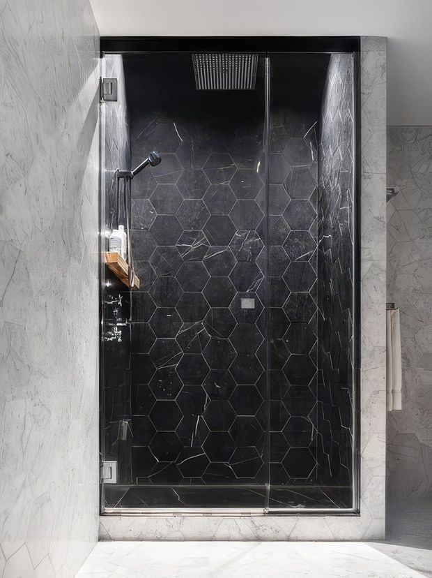 A contemporary walk-in shower enclosure viewed straight on. The shower has a frameless clear-glass door with dark metal edging, a black hexagonal tiled feature wall with white veining, a rainfall showerhead, handheld shower, and recessed shelf. Surrounding walls and floor are finished in pale marble-look material, creating a high-contrast monochromatic bathroom interior.