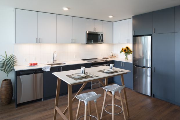 Bright contemporary kitchen with a compact island and integrated dining table, two-tone white and dark cabinetry, stainless-steel appliances, white tiled splashback, and dark timber flooring. The space includes bar stools, recessed ceiling lighting, and simple decorative accents.