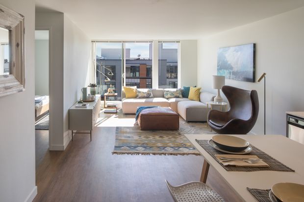 Bright open-plan apartment interior combining a living room and dining area. The room features large windows, light neutral walls, dark timber flooring, a pale sectional sofa with colourful cushions, a sculptural dark leather lounge chair, patterned rug, and a contemporary dining table in the foreground. Urban buildings are visible through the glazing.