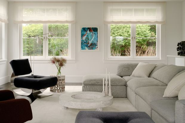 Bright, airy living room with large white-framed windows, light neutral walls and flooring, a pale grey sectional sofa, black lounge chair and ottoman, round stone coffee table, and minimal decorative furnishings. Greenery is visible outside the windows, while a colourful abstract artwork provides a small accent.