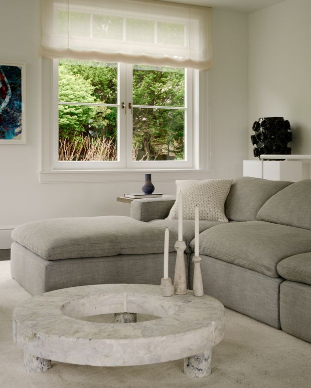 A calm, softly lit living room with a large grey upholstered sectional sofa and chaise, a low round pale stone coffee table, and a white console displaying a dark sculptural object. A double-hung white-framed window with a light Roman blind overlooks leafy greenery, while neutral walls, carpet, cushions, and understated decor create a refined, monochromatic interior.