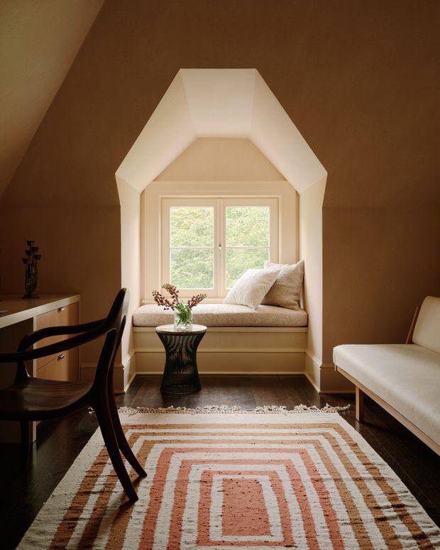 Warm, refined sitting room with a prominent built-in window seat set within a white-trimmed dormer-like alcove. The room has deep brown walls, dark timber flooring, a patterned geometric area rug, a dark wood desk and chair, a cream upholstered bench, and a small decorative side table. Natural light enters through the central divided window, overlooking greenery.
