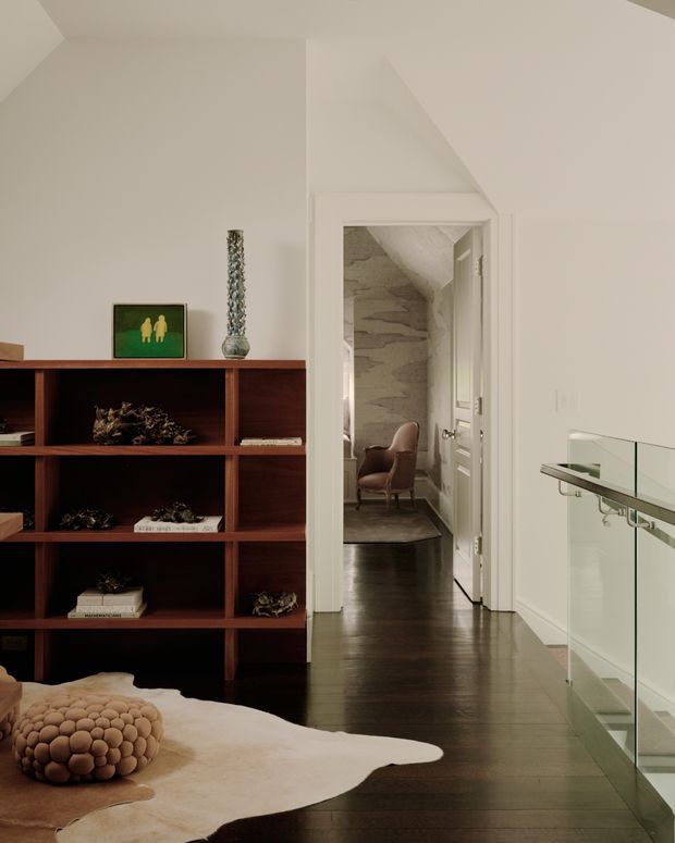 Interior view of a refined modern living area or landing with dark timber built-in shelving, pale walls, dark timber flooring, and a glass balustrade along the right side. A doorway leads to a small adjoining seating area with a sculptural textured stone wall and leather chair. The foreground includes a low upholstered surface with a rounded cushion and a pale hide rug.