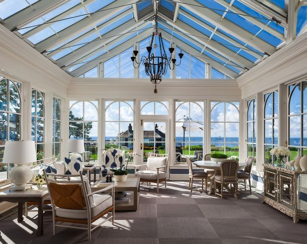 A bright, elegant sunroom or conservatory with extensive white-framed glazing and a fully glazed pitched roof. The room contains upholstered lounge seating, occasional tables, a dining area, decorative cabinetry, plants, and a central black chandelier. Ocean views are visible through the windows, while the interior is finished in white trim with patterned neutral carpeting and refined coastal furnishings.