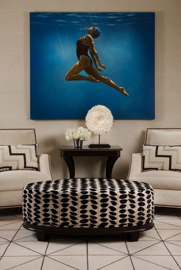 A refined living room with two cream upholstered armchairs facing a large oval ottoman upholstered in a bold black-and-cream leaf pattern. A dark wood console table sits between the chairs, topped with flowers and a sculptural lamp, while a large blue underwater artwork forms the focal point above. The room has light tiled flooring with geometric dark grout lines and a formal, high-contrast decorative scheme.