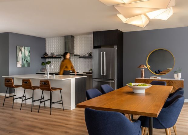 Open-plan kitchen and dining area with a contemporary dark grey and timber palette. The kitchen features a white island with integrated sink and bar seating, dark cabinetry, stainless-steel refrigerator, and a textured white tiled splashback. A large timber dining table with navy upholstered chairs occupies the foreground, while a geometric sculptural ceiling light, circular gold-framed mirror, and compact sideboard add decorative accents.