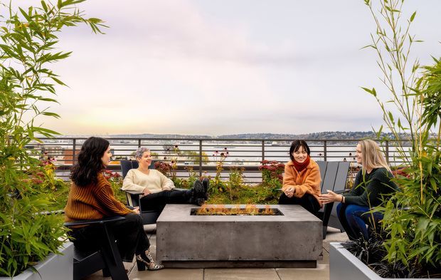 A landscaped rooftop terrace or balcony with four people seated around a large rectangular concrete fire pit or feature table. Tall bamboo planters frame the outdoor lounge, with metal balustrading and a broad waterfront or cityscape view beyond. The scene has a relaxed, contemporary outdoor entertaining character.