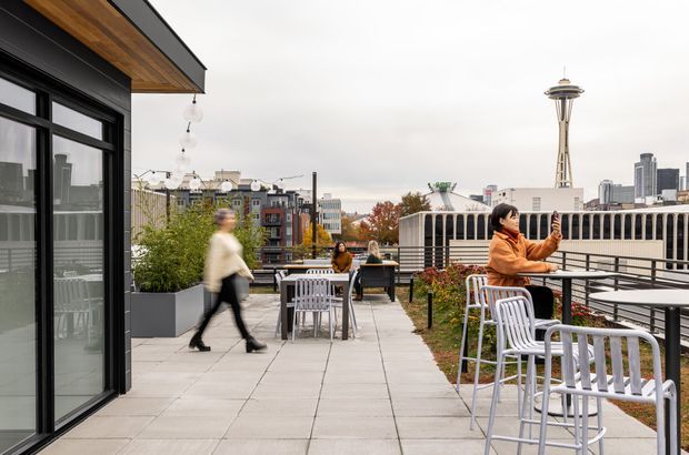 A contemporary rooftop terrace with paved outdoor seating, high tables and stools, large planters, and a glass-walled building edge. The terrace overlooks an urban skyline, with the Space Needle prominently visible in the background. The image has an overcast, urban atmosphere and shows a mix of black-framed glazing, warm timber soffit lining, concrete paving, and white metal furniture.