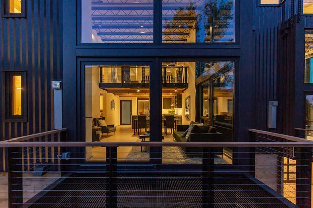 Exterior dusk view of a dark-clad contemporary multi-storey residence, focused on a glazed balcony or terrace overlooking a double-height interior living area. Large black-framed windows reveal warm timber-toned floors, seating, dining, kitchen cabinetry and an upper-level walkway inside. The facade has vertical dark cladding, narrow windows, exposed balcony structure and horizontal metal cable railings.