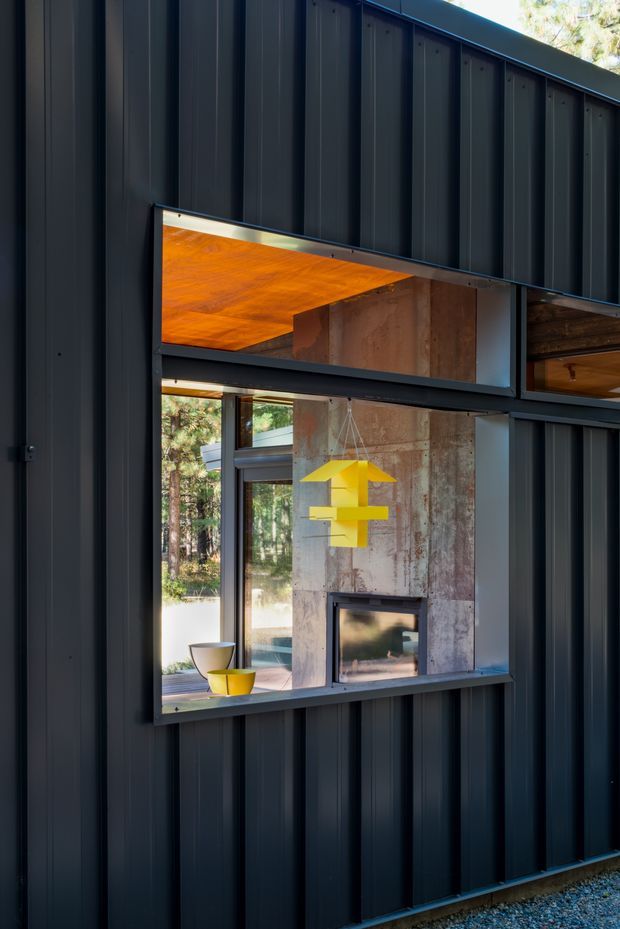 Exterior view of a contemporary dark-clad building with a projecting, deeply framed window opening revealing a living room fireplace, concrete feature wall, timber ceiling, and bright yellow pendant light inside. The facade uses vertically ribbed dark metal cladding and angular roof geometry, with forest visible beyond the glazing.